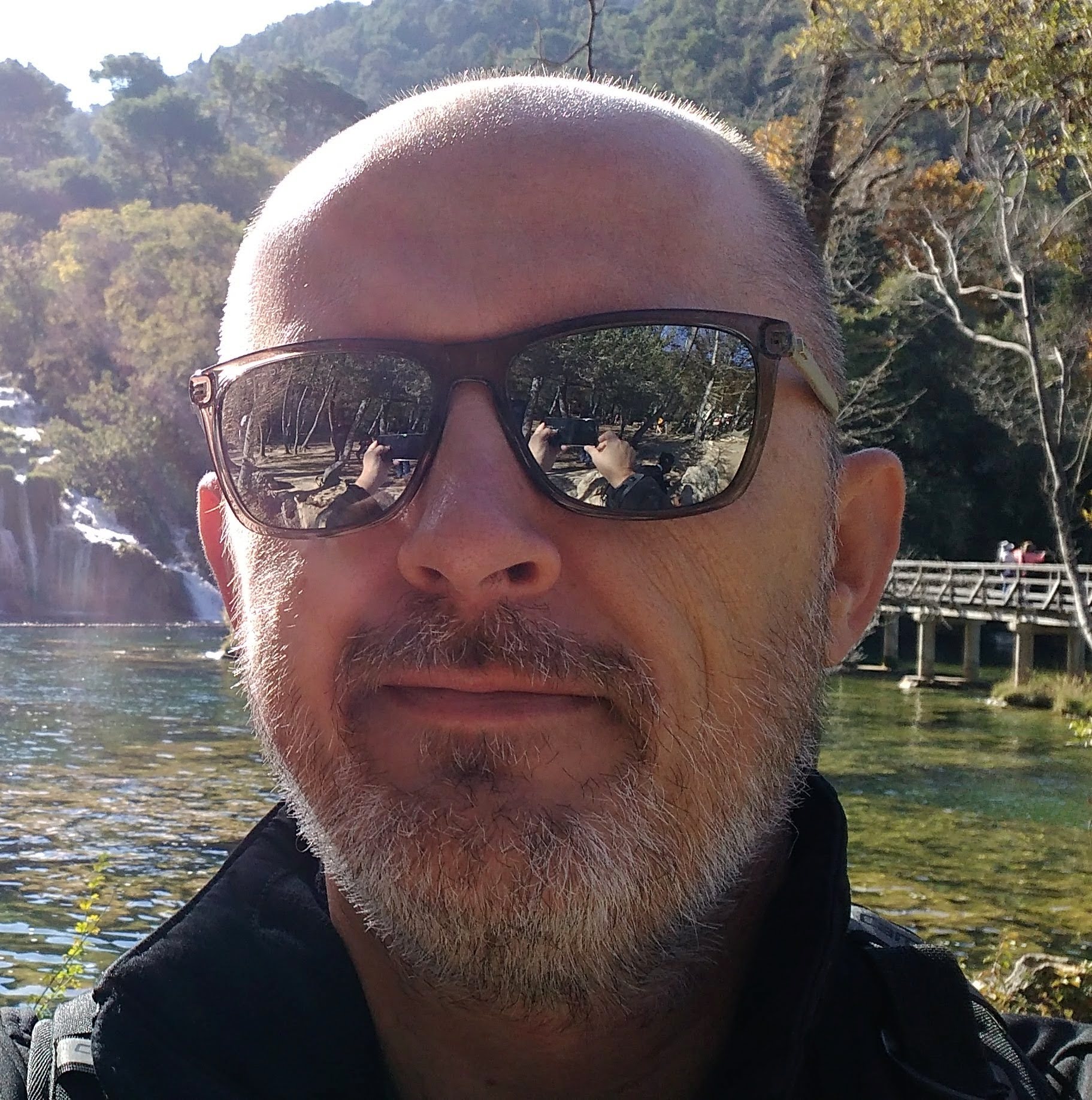 Zdjęcie prowadzącego webinar – Bartosza Krupy. Mężczyzna w okularach przeciwsłonecznych nad jeziorem z wodospadem w tle.